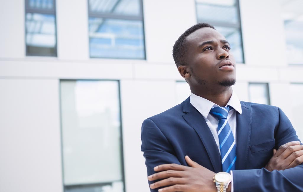 Un giovane professionista in un abito blu navy con cravatta a righe sta con sicurezza le braccia incrociate, guardando in lontananza davanti a un edificio moderno, rappresentando leadership, successo aziendale e formazione manageriale.